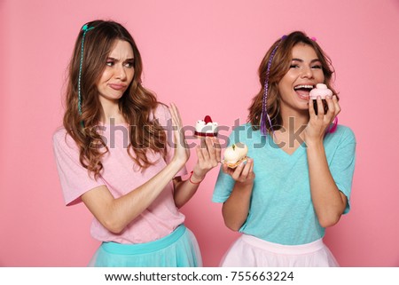Portrait of two doubtful smiling girls in colorful bright clothes holding cupcakes isolated over pink background