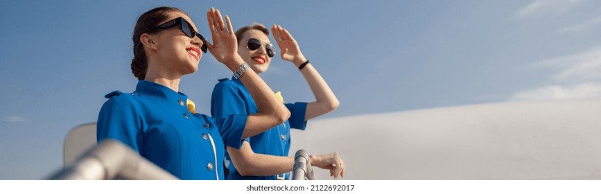 Portrait of two elegant air stewardesses in blue uniform and sunglasses covering eyes with hand and looking far away, standing together on airstair. Aircrew, occupation concept - Shutterstock ID 2122692017