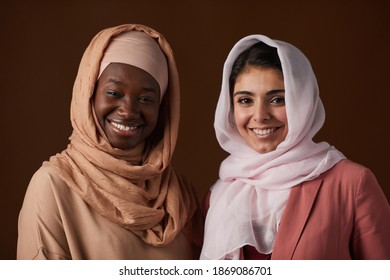 Portrait of two ethnic young women wearing headscarves and smiling at camera while posing against brown background in studio and celebrating heritage and personal identity