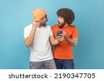 Portrait of two excited amazed young adult hipster men standing with cell phone and celebrating success, clenched fists, looking at each other. Indoor studio shot isolated on blue background.