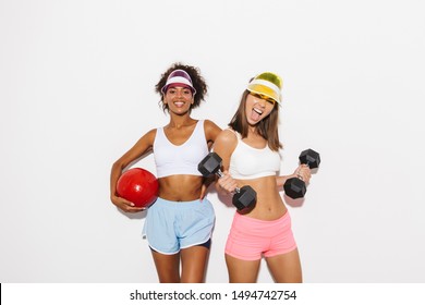 Portrait Of Two Excited Multiethnic Women Wearing Visor Hats Poising With Fitness Ball And Dumbbells Isolated Over White Background