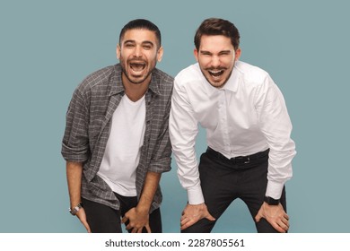 Portrait of two extremely happy laughing men friends, looking at camera, see something very funny, keeping mouth opened. Indoor studio shot isolated on light blue background.