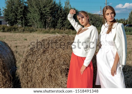 Portrait of two fair-haired girls in fashionable and stylish clothes, against the background of a field and a stack of straw. On a sunny day in August.