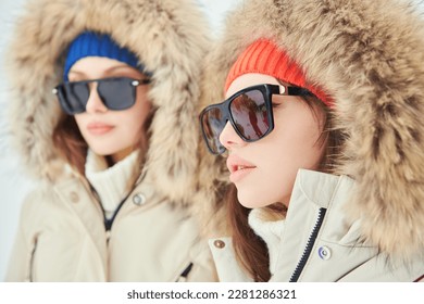 Portrait of two fashion model girls posing in white downy overalls against outdoors. Alpine skiing, active winter recreation. Winter fashion.
