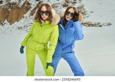Portrait of two fashionable girls in bright downy overalls and trendy sunglasses posing against a snowy winter background. Winter fashion. Vacation in a ski resort.