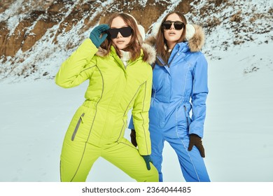 Portrait of two fashionable girls in bright downy overalls and trendy sunglasses posing against a snowy winter background. Winter fashion. Vacation in a ski resort.