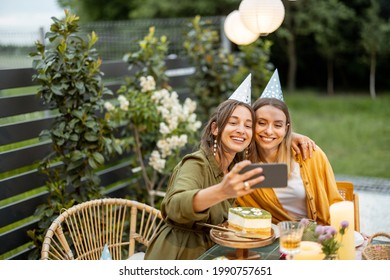 Portrait Of A Two Female Friends Celebrating Birthday, Making Selfie Or Callin On Phone While Sitting By A Festive Table At Backyard Outdoors