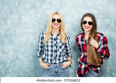 Portrait of two female friends in checkered shirts, jeans and sunglasses on the blue painted wall background