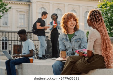 Portrait of two female multicultural university students sitting on stairs at college campus on a coffee break and smiling at each other while chatting. In background are diverse students studying. - Powered by Shutterstock