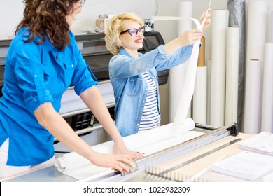 Portrait of two female operators in modern printing shop or publishing company, focus on smiling blonde woman cutting  paper and loading plotter machines, copy space