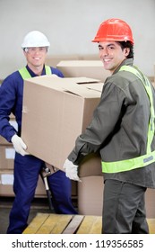 Portrait Of Two Foremen Lifting Cardboard Box In Warehouse