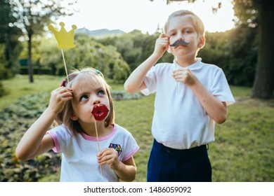 Portrait of two funny kids playing together with paper accessories on a stick for photo shoot on holiday or party in autumn park. Brother sister holds paper lips, crown, mustache, hat. Children's day