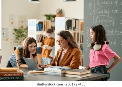 Portrait of two girls talking to female teacher using laptop during consultation in school library - Powered by Shutterstock