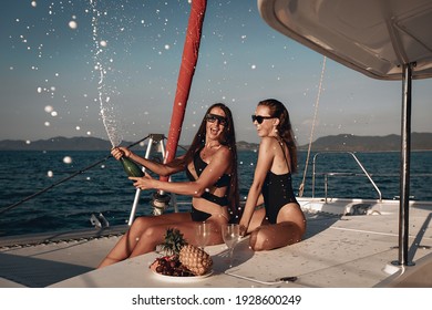 Portrait of two gorgeous radiant girlfriends celebrating the holiday by opening champagne. The champagne spray is everywhere. Two girls are having fun on their huge white yacht. Fun concept