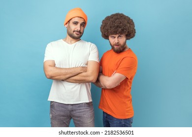 Portrait Of Two Handsome Serious Young Adult Hipster Men Standing Looking At Camera With Assertive Expression, Keeping Hands Folded. Indoor Studio Shot Isolated On Blue Background.