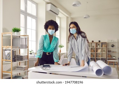 Portrait Of Two Happy Architects, Designers Or Business Women In Face Masks Standing In Office Studio. Return To Workplace After End Of Lockdown And Quarantine. Safety During Coronavirus Pandemic