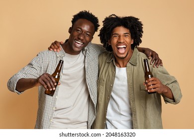 Portrait Of Two Happy Black Friends Embracing And Holding Bottles With Beer, Cheerful African American Guys Having Fun While Posing Together Over Beige Background In Studio, Enjoying Party