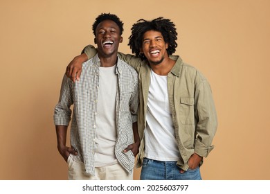 Portrait Of Two Happy Black Guys Embracing While Posing Over Beige Background, Cheerful Young African American Friends In Stylish Clothes Having Fun In Studio, Laughing At Camera, Copy Space