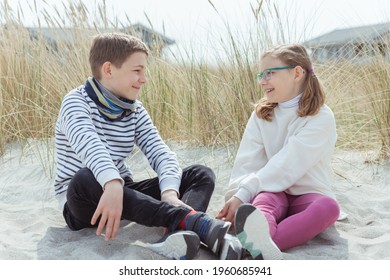 Portrait Of Two Happy Cute Teen Siblings Sitting And Joyful Talking On White Sand At Baltic Sea Beach With Dunes