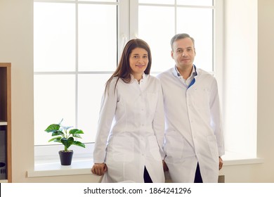 Portrait Of Two Happy Male And Female Doctors In Medical Uniform Standing By The Window And Smiling Looking At Camera. Concept Of Professional Medicine And A Strong Medical Team.