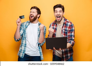 Portrait Of A Two Happy Young Men Holding Laptop Computer While Standing With Credit Card And Celebrating Isolated Over Yellow Background