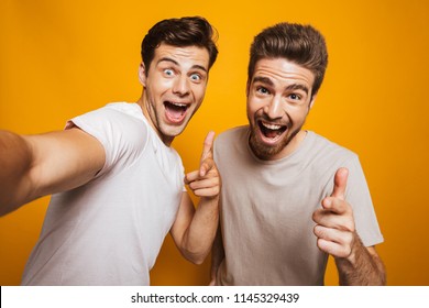 Portrait Of A Two Happy Young Men Best Friends Pointing Fingers At Camera Taking A Selfie Isolated Over Yellow Background