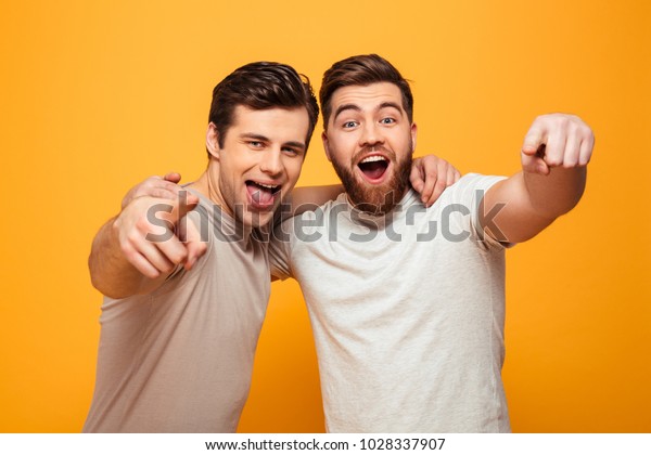 黄色い背景にカメラを指差す2人の幸せな若い男性のポートレート の写真素材 今すぐ編集