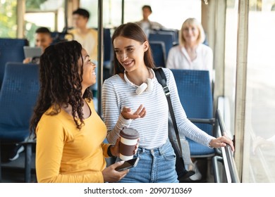 Portrait of two happy young multiethnic ladies standing inside bus and chatting, drinking coffee from cup and using cellphone technology, friends commuting together to work or high school university - Powered by Shutterstock