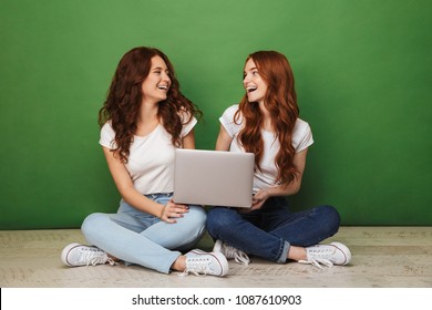 Portrait Of Two Happy Young Redhead Girls Sitting On A Floor With Laptop Computer And Looking At Each Other Isolated Over Green Background