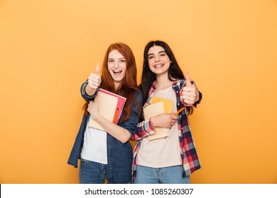 Portrait of two happy young school teenage girls holding copybooks and showing thumbs up isolated over yellow background