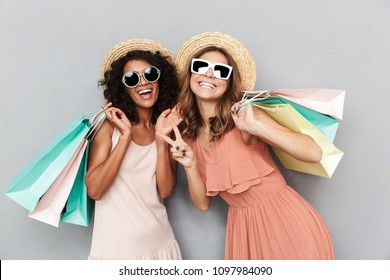 Portrait of two happy young women dressed in summer clothes holding shopping bags and showing peace gesture isolated over gray background