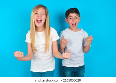Portrait of two kids boy and girl standing over blue background looks with excitement at camera, keeps hands raised over head, notices something unexpected reacts on sudden news.