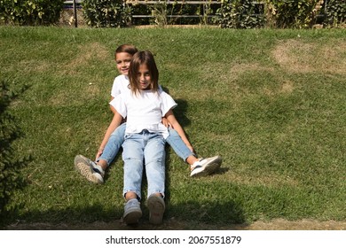 Portrait Of Two Little Brother And Sister Children Lying On The Grass. They Are Dressed In White T-shirts And Jeans. The Older Sister Is A Trans Girl. Concept Of A United And Happy Family.