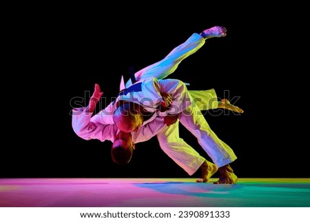 Portrait of two male sportsmen, martial arts fighters in white kimono fighting performing techniques in motion in neon light isolated black background. Concept of combat sport, energy. Copy space