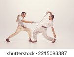 Portrait of two men in a suit fighting with swords isolated over grey studio background. Concept of sport, hobby, emotions, active lifestyle, retro fashion. Copy space for ad