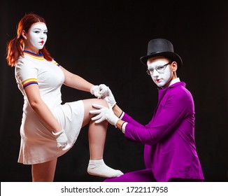 Portrait of two mime artists, isolated on black background. Young woman puts her foot on mans knee. Symbol of worship, mistress, subordination