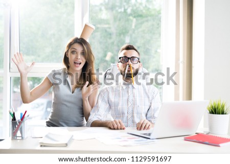 Portrait of two modern business people goofing around having fun while looking at camera sitting at desk in sunlit office