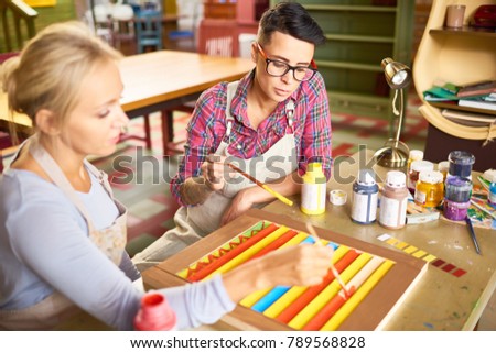 Portrait of two modern female artists enjoying  working together in art studio painting together sitting at wooden desk