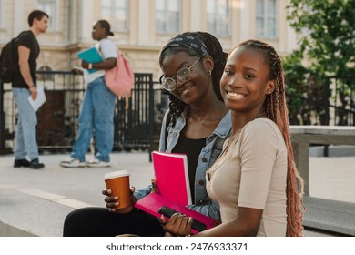 Portrait of two multicultural happy university student girls sitting on stairs at college campus with textbooks and coffee in hands and smiling at the camera. Young diverse students posing at campus. - Powered by Shutterstock