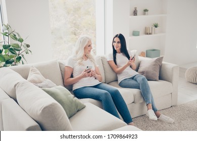Portrait of two nice attractive lovely cheerful cheery women sitting on divan using digital device web app spending free time day weekend in white light interior house flat apartment