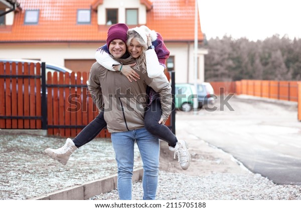 Portrait of two\
people couple laughing smiling fair-haired woman and man together\
jump riding outdoors in frost weather near house. Cars houses and\
fence on background. Side\
view