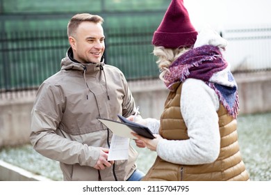 Portrait Of Two People Smile Man And Blonde Hair Woman Back With Documents Holding Pencil Near House. Speaking To Each Other And Deal Sign Documents And Calculate. Together. Side View Light Day.