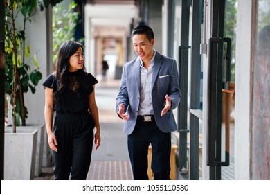 A Portrait Of Two Professional Business People Walking And Talking. They Are Deep In Conversation As They Walk On A Street In A City In Asia. The Man And Woman Are Both Professionally Dressed. 