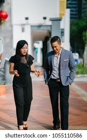 A Portrait Of Two Professional Business People Walking And Talking. They Are Deep In Conversation As They Walk On A Street In A City In Asia. The Man And Woman Are Both Professionally Dressed. 