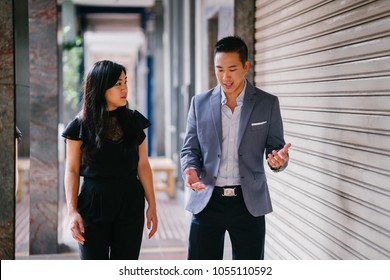 A Portrait Of Two Professional Business People Walking And Talking. They Are Deep In Conversation As They Walk On A Street In A City In Asia. The Man And Woman Are Both Professionally Dressed. 