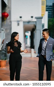 A Portrait Of Two Professional Business People Walking And Talking. They Are Deep In Conversation As They Walk On A Street In A City In Asia. The Man And Woman Are Both Professionally Dressed. 