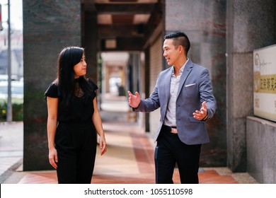 A Portrait Of Two Professional Business People Walking And Talking. They Are Deep In Conversation As They Walk On A Street In A City In Asia. The Man And Woman Are Both Professionally Dressed. 