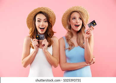 Portrait of a two satisfied girls dressed in swimsuits and summer hats holding credit cards isolated over pink background