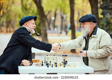 Portrait of two senior men playing chess in the park on a daytime in fall. Good game. Shaking hands. Concept of leisure activity, friendship, sport, autumn season, game, entertainment, old generation - Shutterstock ID 2230598123