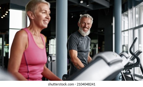 Portrait Of Two Senior People Working Out On Elliptical Machine In Gym. Selective Focus Of Smiling Grey-haired And Bearded Man Looking At Blond Sportive Woman. Seniors Workout In Gym.